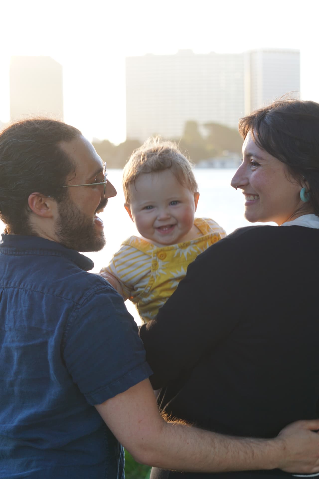 The Espicha-Blumenstein parents holding their laughing baby between them in golden hour light at Lake Merritt