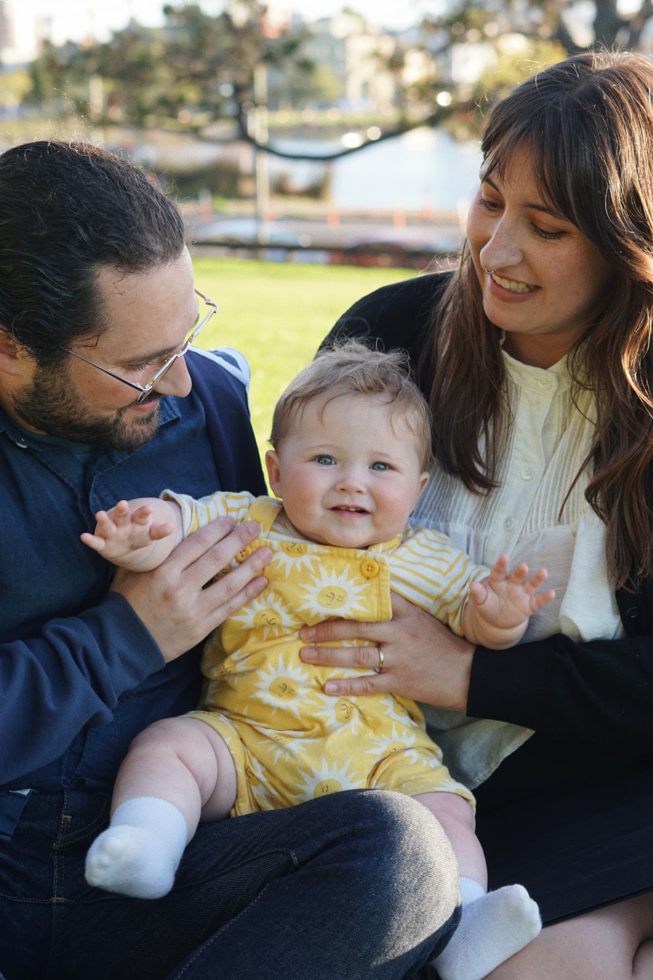 The Espicha-Blumenstein parents smiling down at their baby seated on their laps in the park