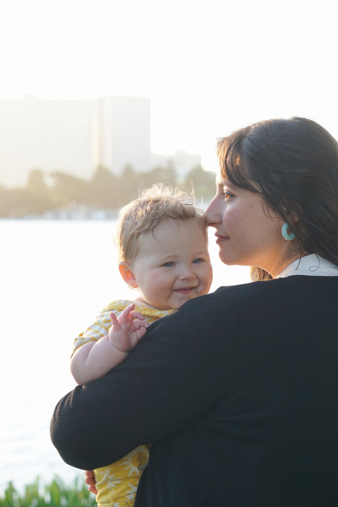 A mother holding her smiling baby close by the water at Lake Merritt in backlit golden light