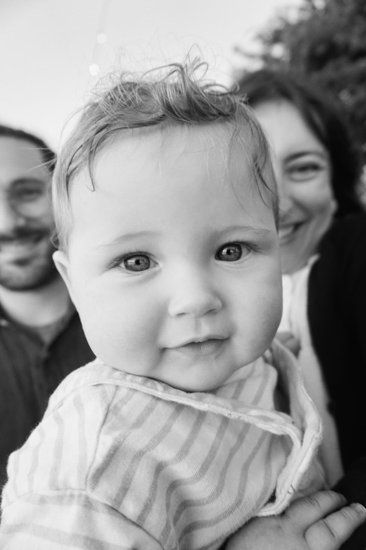 Black and white close-up of the Espicha-Blumenstein baby smiling with her parents blurred behind her