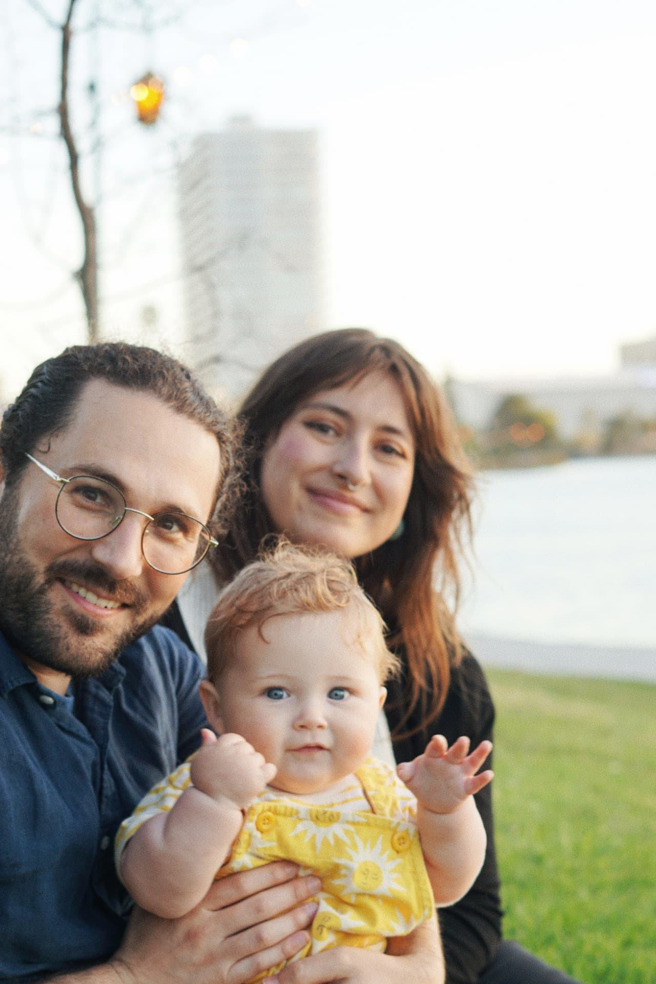 The Espicha-Blumenstein parents holding their baby in a sunny outfit by Lake Merritt at sunset