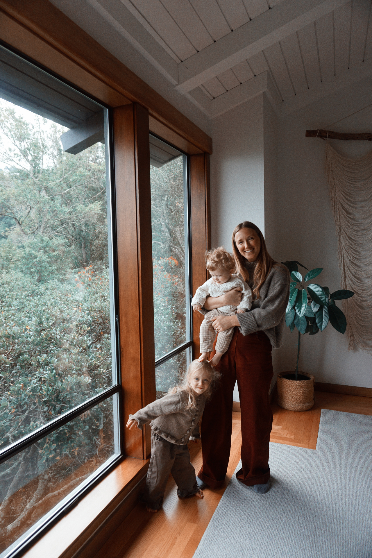 The Fleischmann mother holding her baby beside her daughter by tall wooden windows overlooking the trees