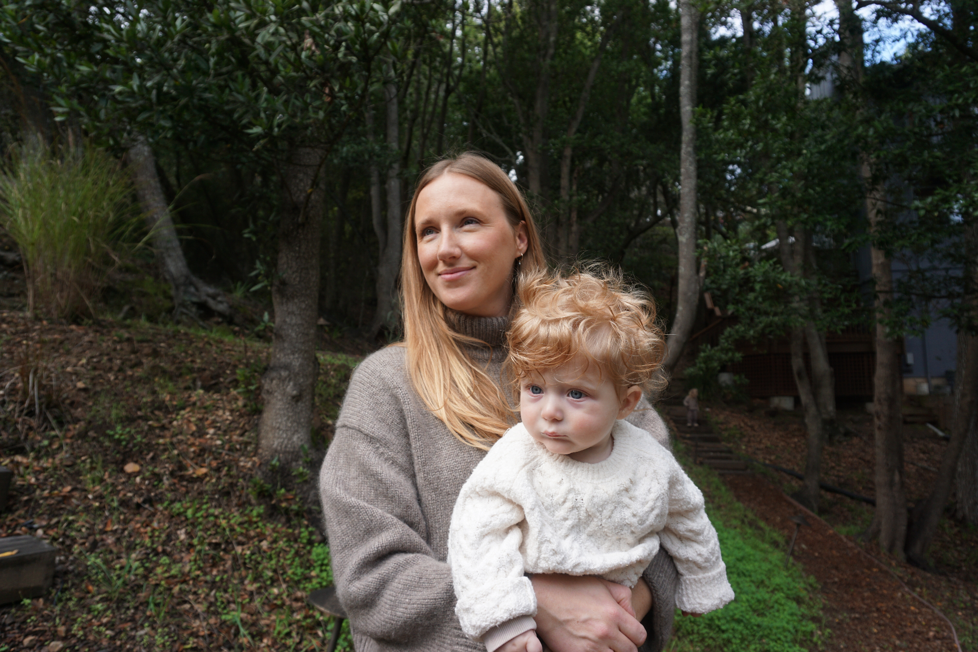 The Fleischmann mother holding her curly-haired toddler outdoors among the trees