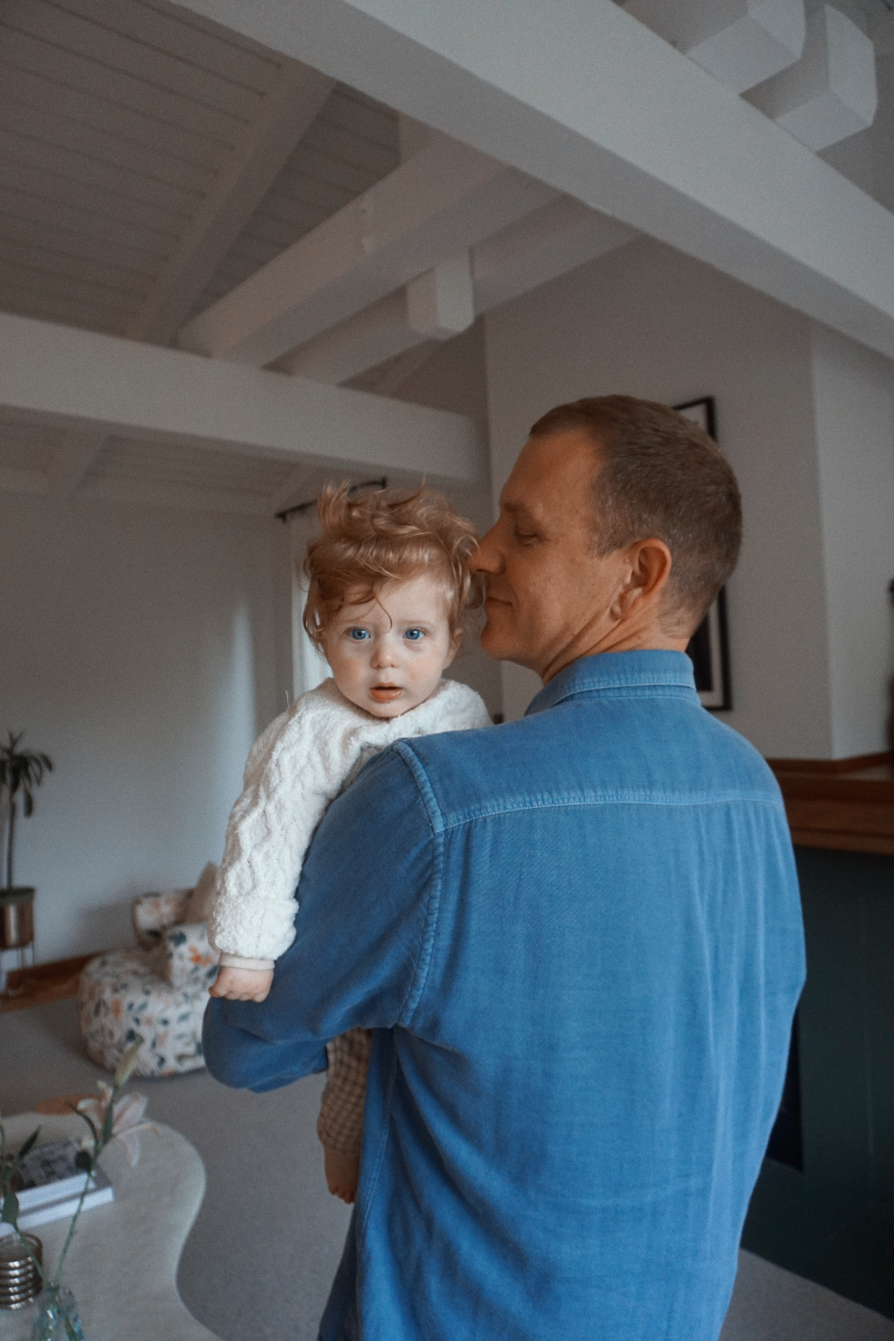 The Fleischmann father holding his curly-haired baby in a softly lit living room with white wood-beamed ceilings