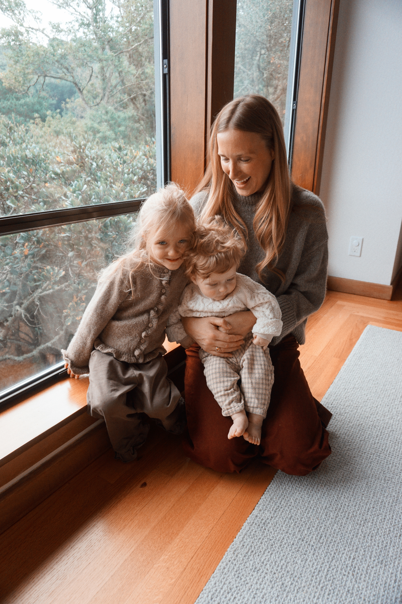 The Fleischmann mother sitting by a large window with her daughter and baby, smiling together with treetops in the background