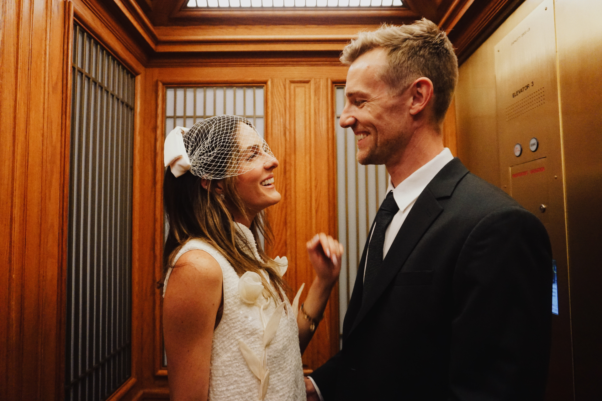 Maggie and Jono smiling at each other inside a wood-paneled San Francisco City Hall elevator after their civil ceremony