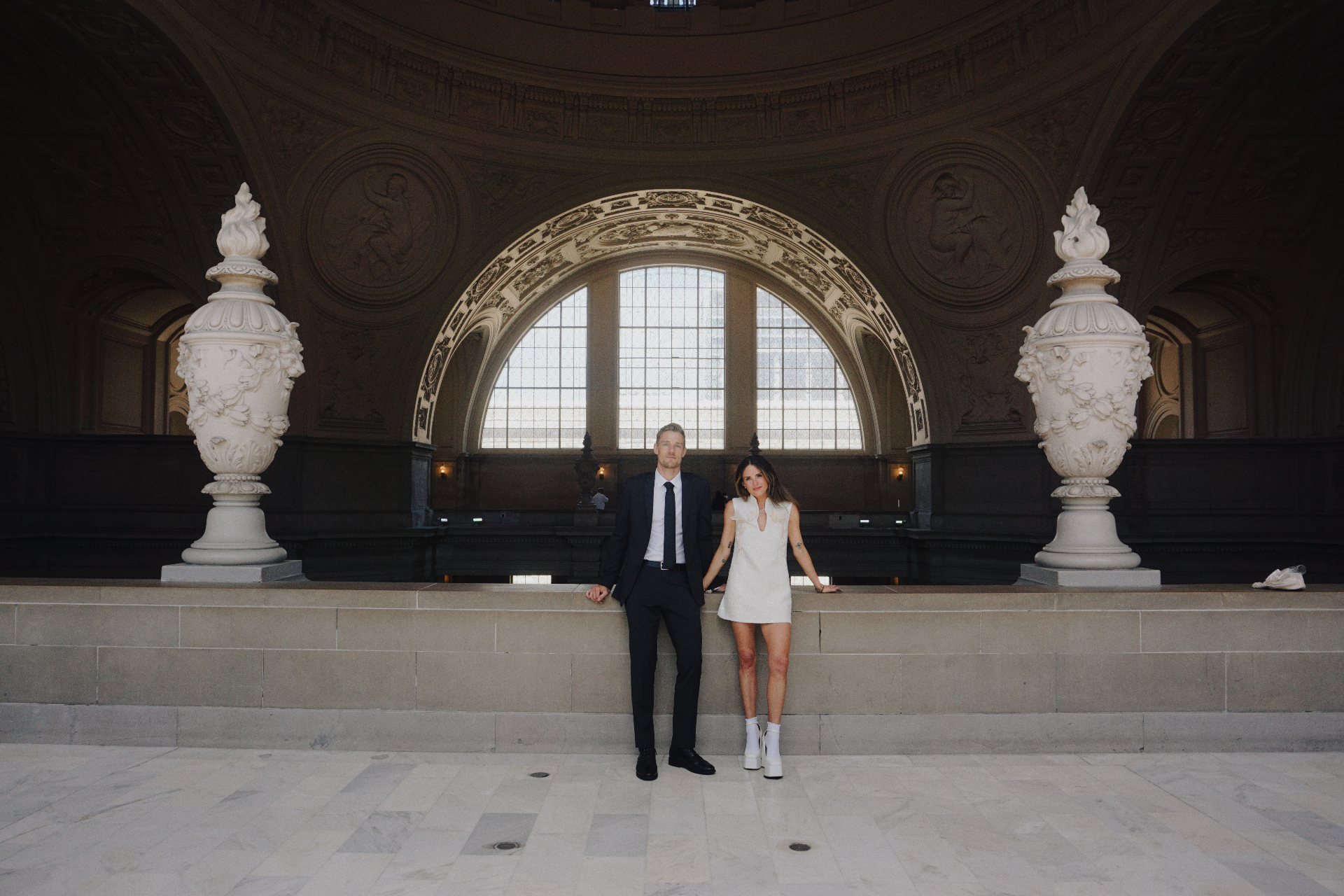 Maggie and Jono standing between ornate urns beneath the grand arched window at San Francisco City Hall