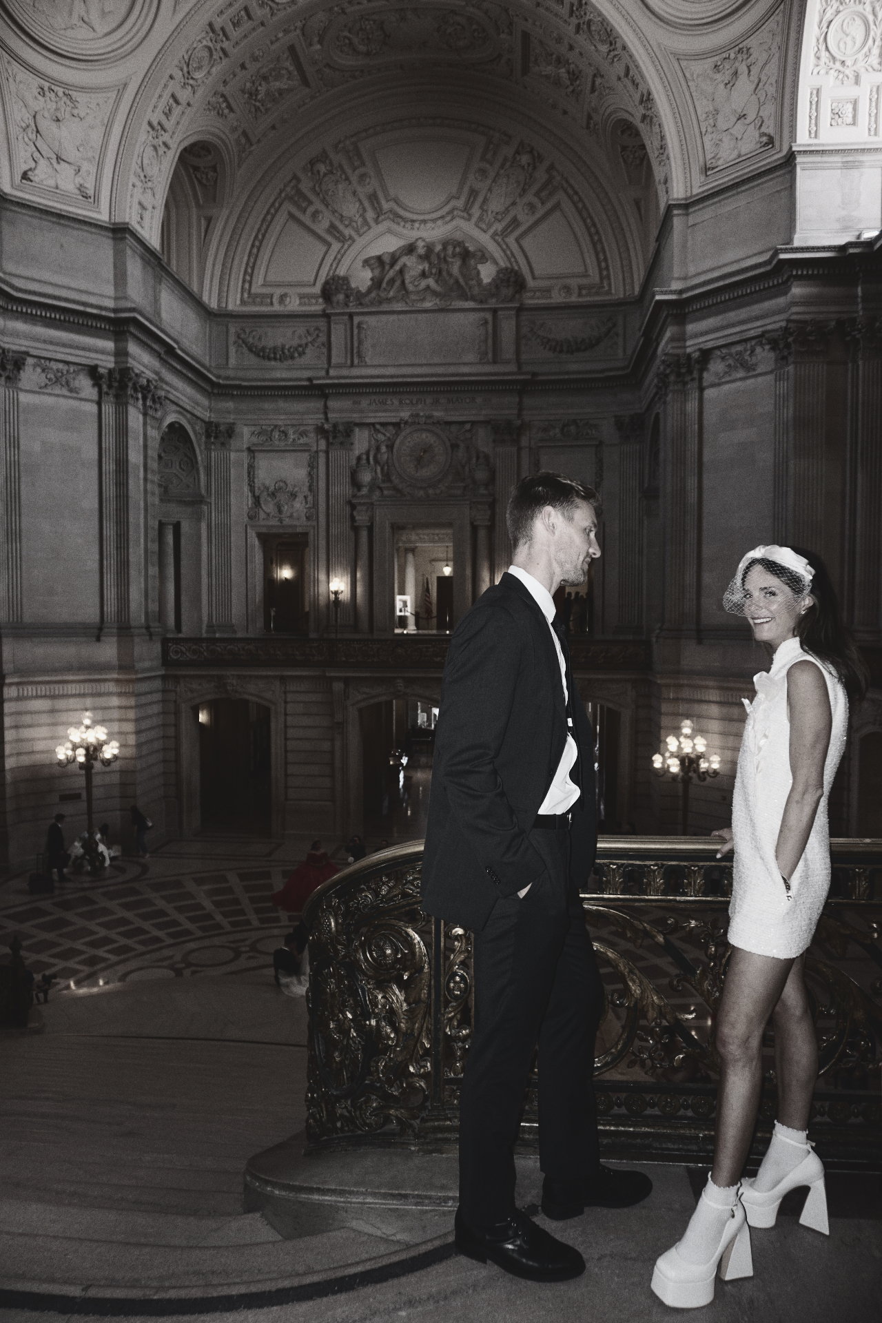 Black and white portrait of Maggie smiling at Jono beside the ornate gilded balustrade beneath the grand rotunda at San Francisco City Hall