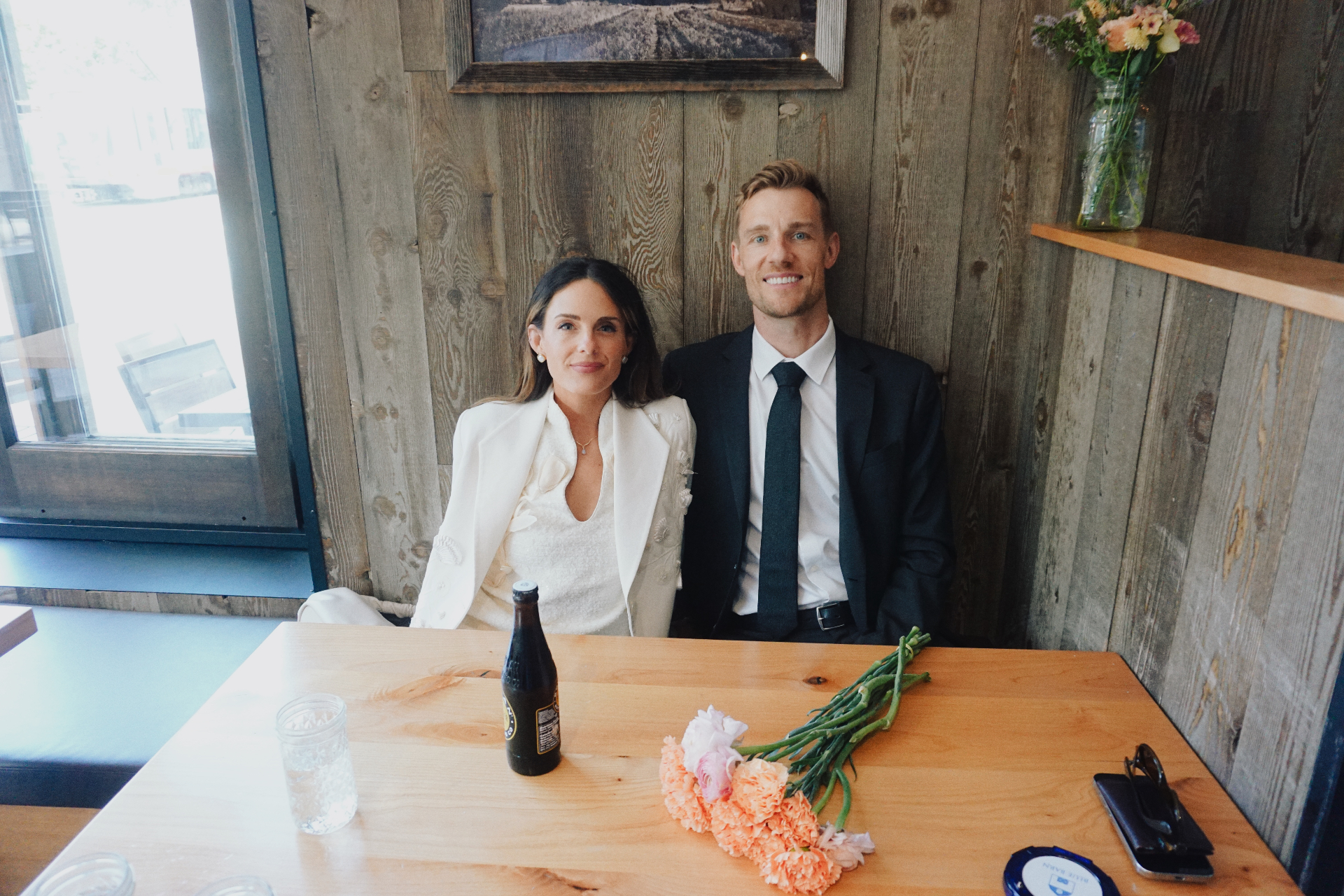 Maggie and Jono sitting together at a wooden booth with flowers and a bottle on the table after their City Hall ceremony