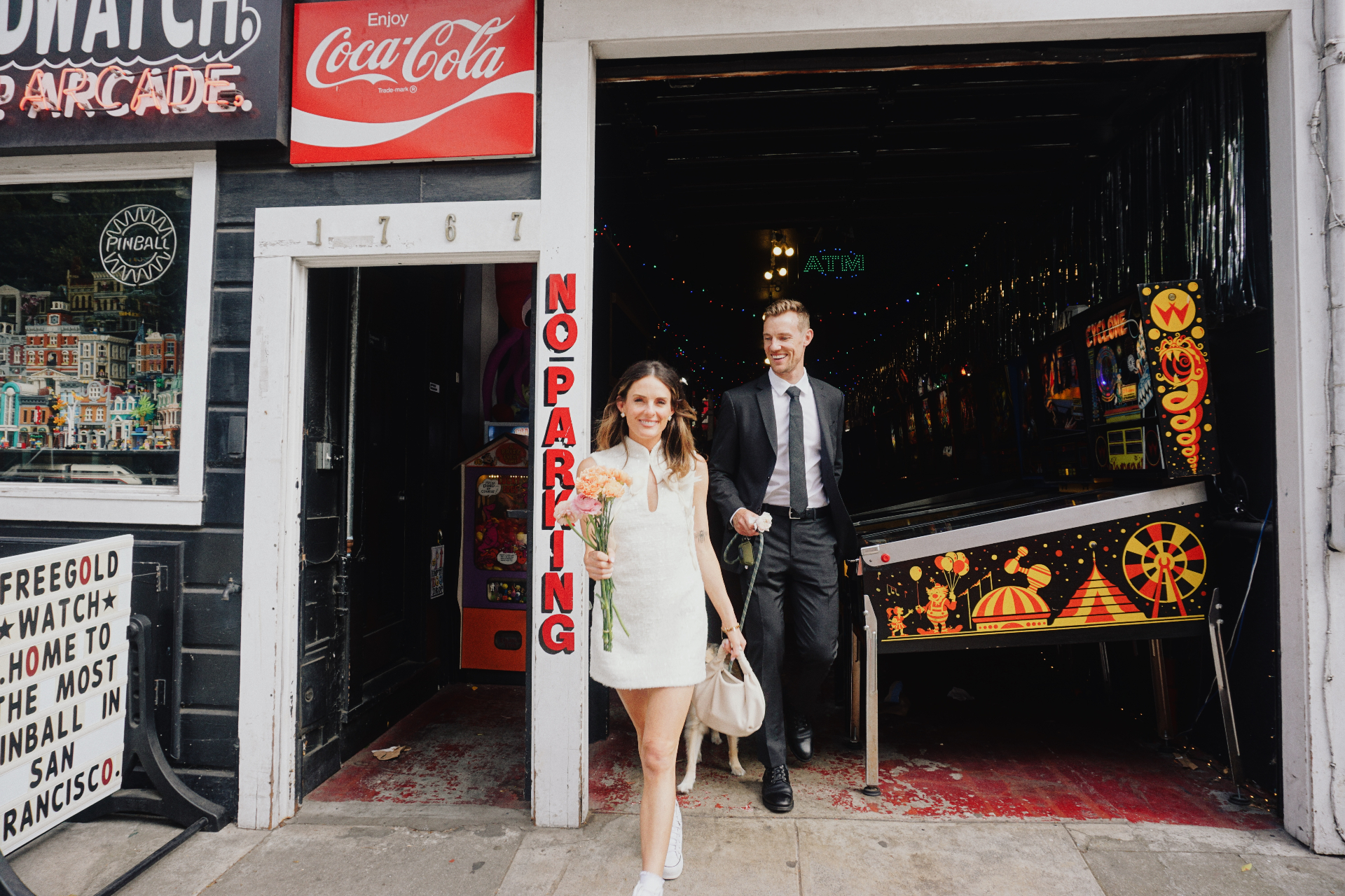 Maggie and Jono smiling as they walk out of a San Francisco pinball arcade with flowers and their dog