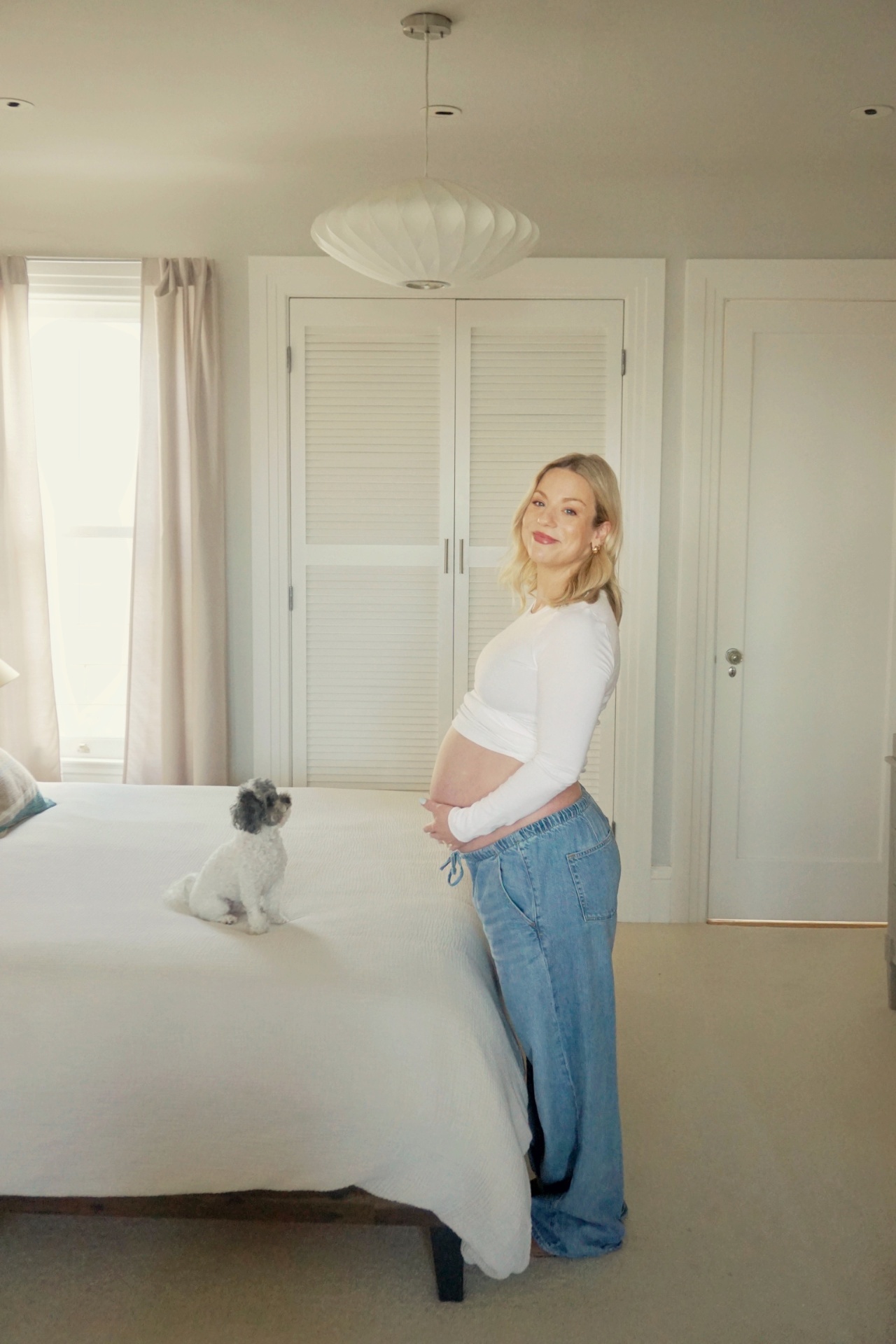 The Parson mother smiling in profile cradling her baby bump in a sunlit bedroom with their small dog on the bed