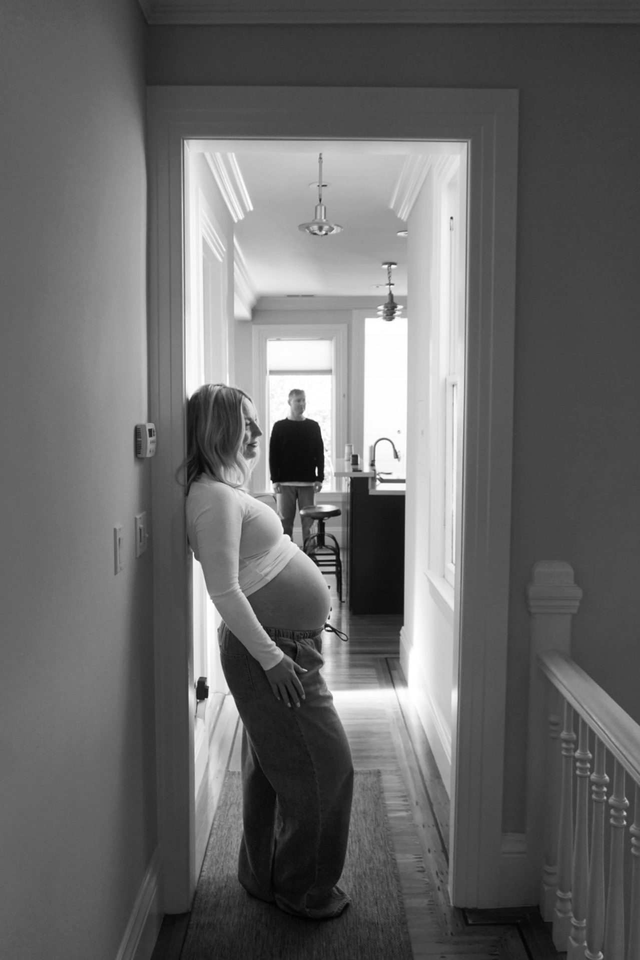 Black and white maternity portrait of The Parson mother leaning in a doorway while her partner stands in the kitchen down the hallway