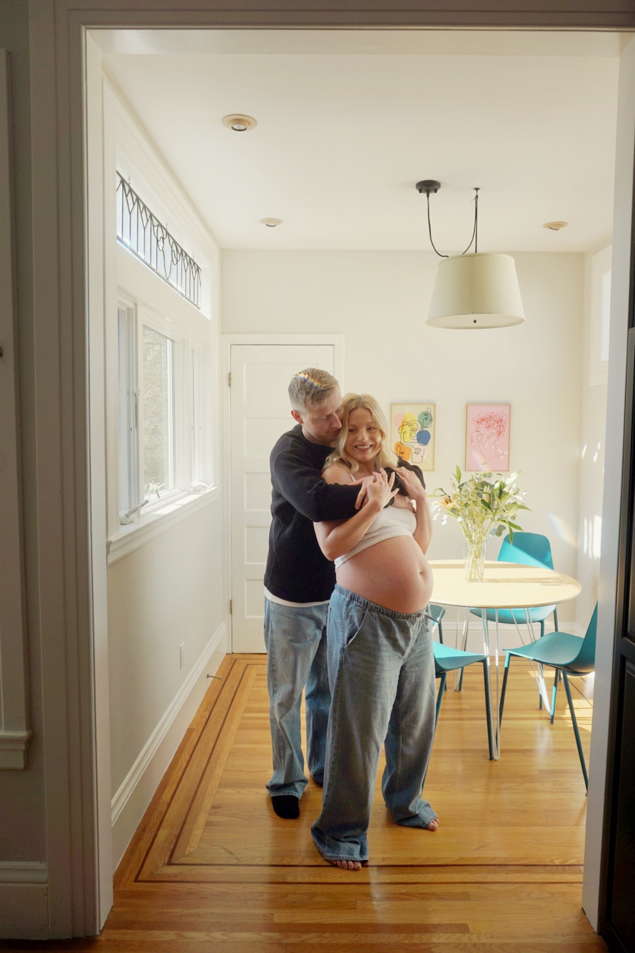 The Parson father embracing the expecting mother from behind in their sunlit dining room
