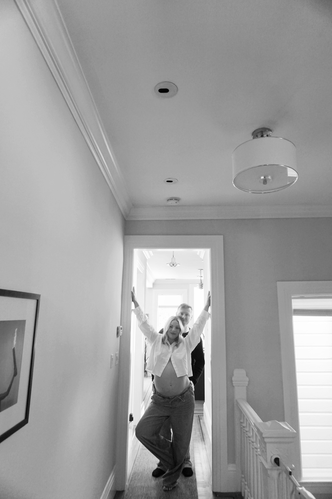 Black and white photo of The Parson mother holding the doorframe with arms raised while her partner stands behind her in the hallway