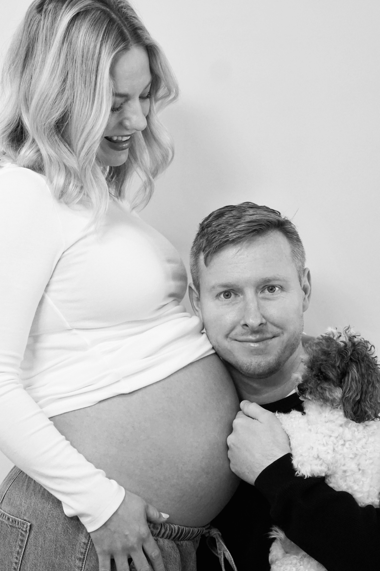Black and white close portrait of The Parson father resting against the mother's baby bump while holding their small dog