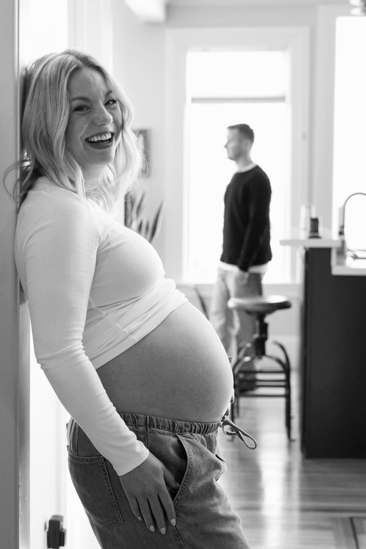 Black and white profile portrait of The Parson mother laughing in the doorway with her partner softly out of focus in the kitchen behind her
