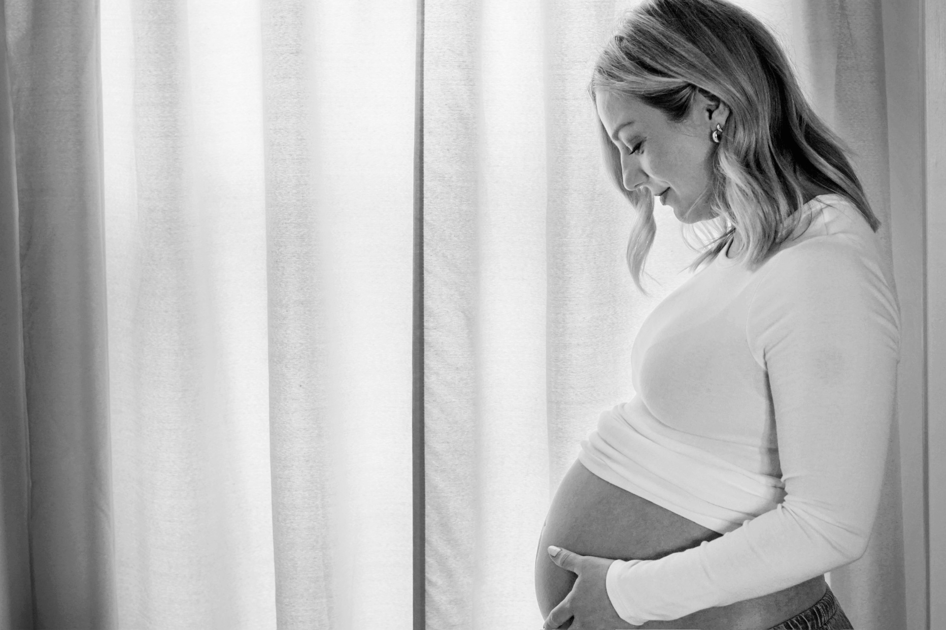 Black and white silhouette of The Parson mother cradling her baby bump in front of soft sunlit curtains