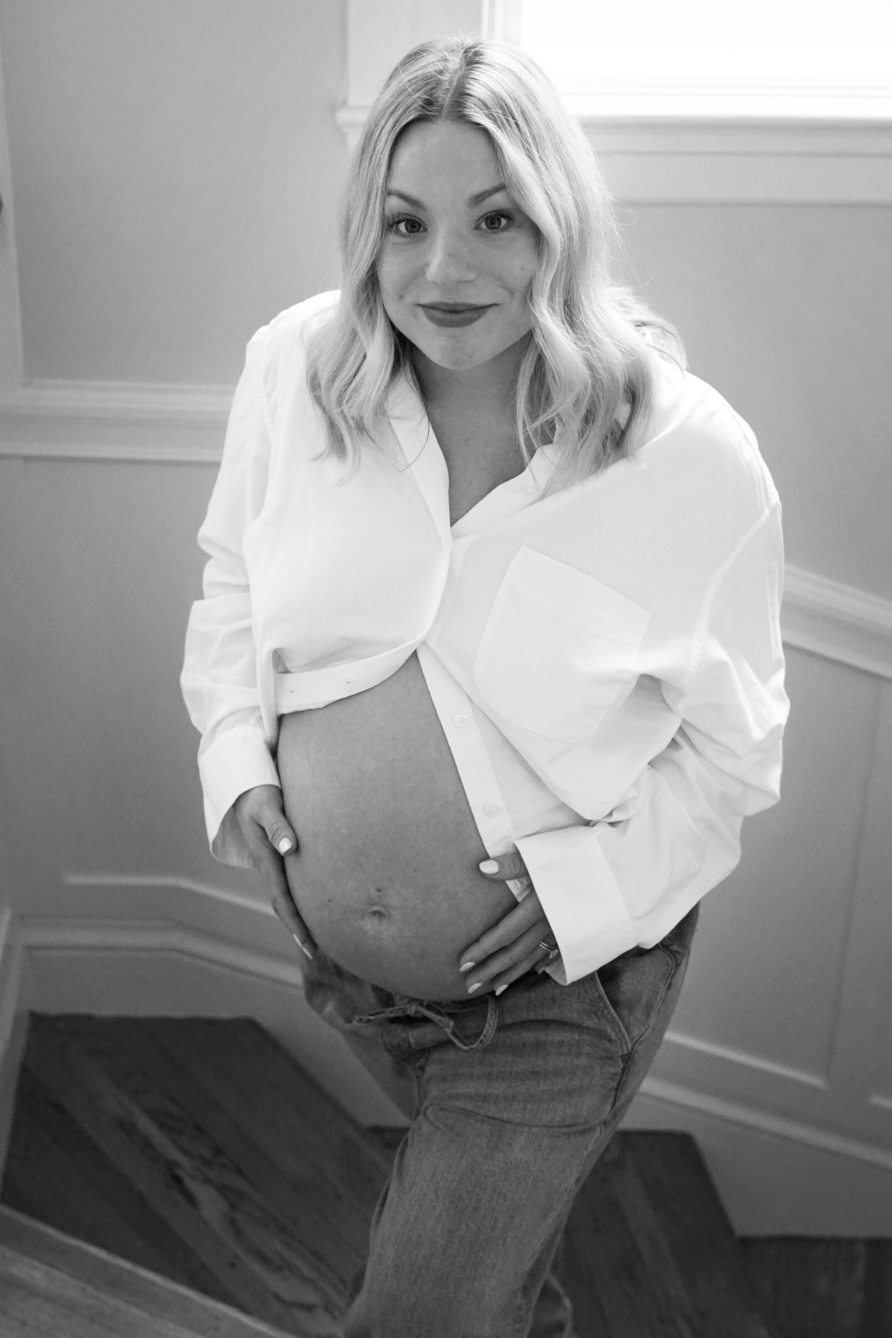 Black and white portrait of The Parson mother in an open white button-down shirt softly holding her baby bump on the staircase
