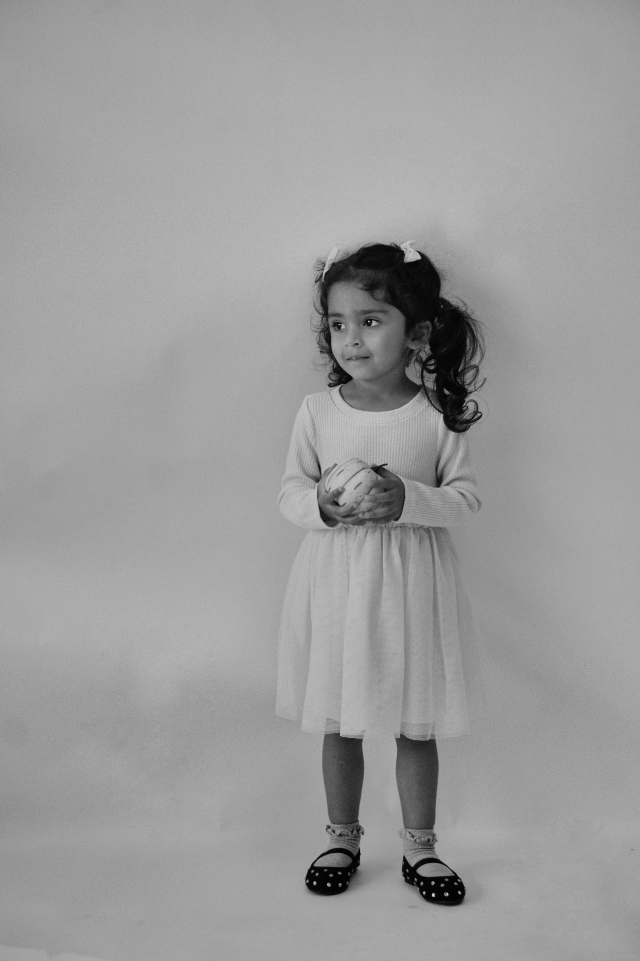 Black and white full-length portrait of The Ramachandran daughter holding a small pumpkin in a tulle dress