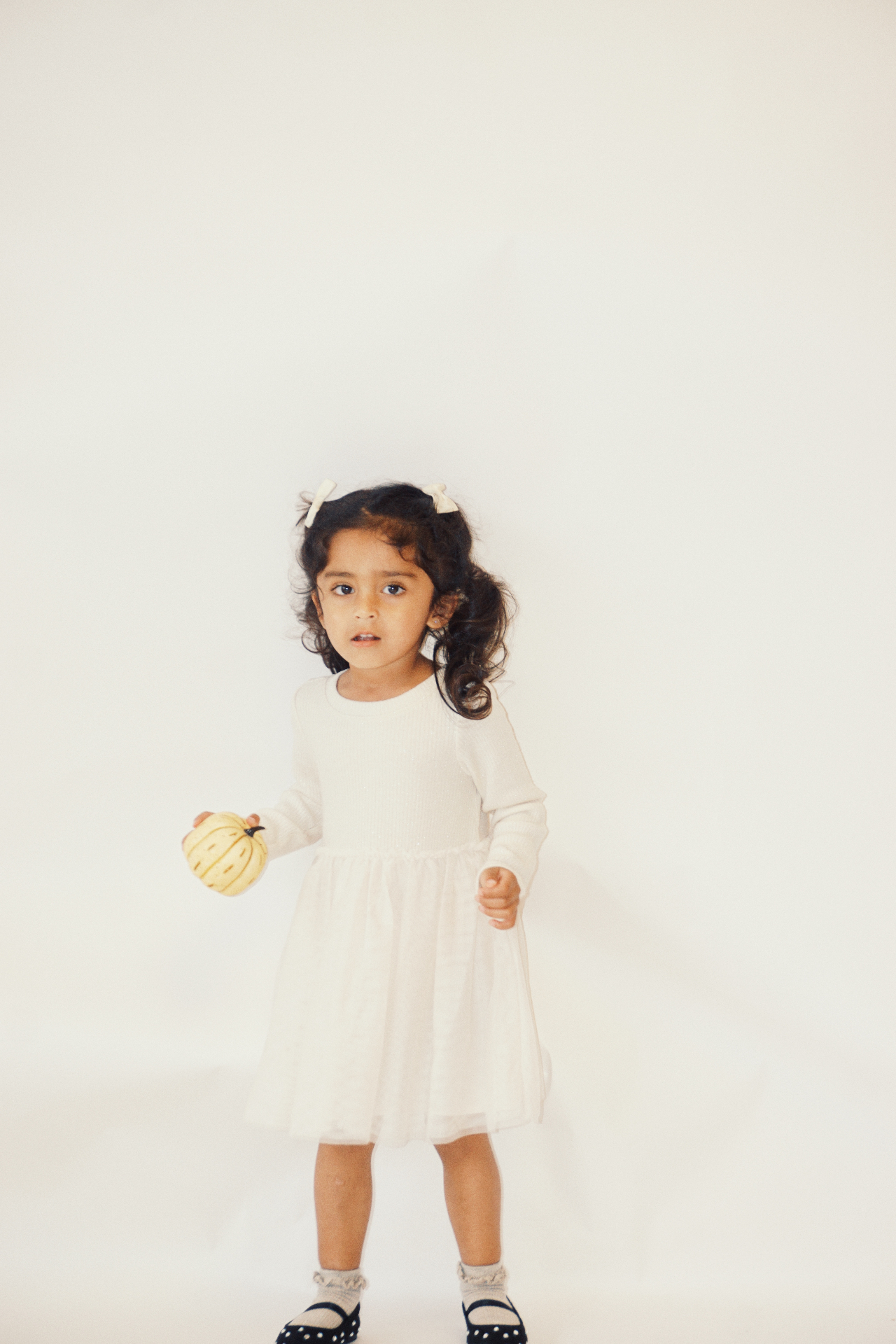 The Ramachandran daughter in a white tulle dress holding a small yellow pumpkin against a soft white backdrop