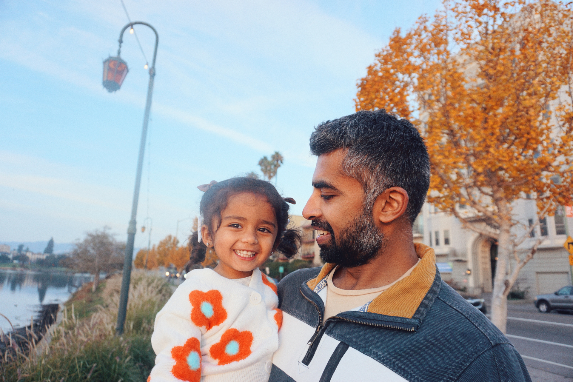 The Ramachandran father smiling at the camera holding his daughter by Lake Merritt with autumn trees behind them