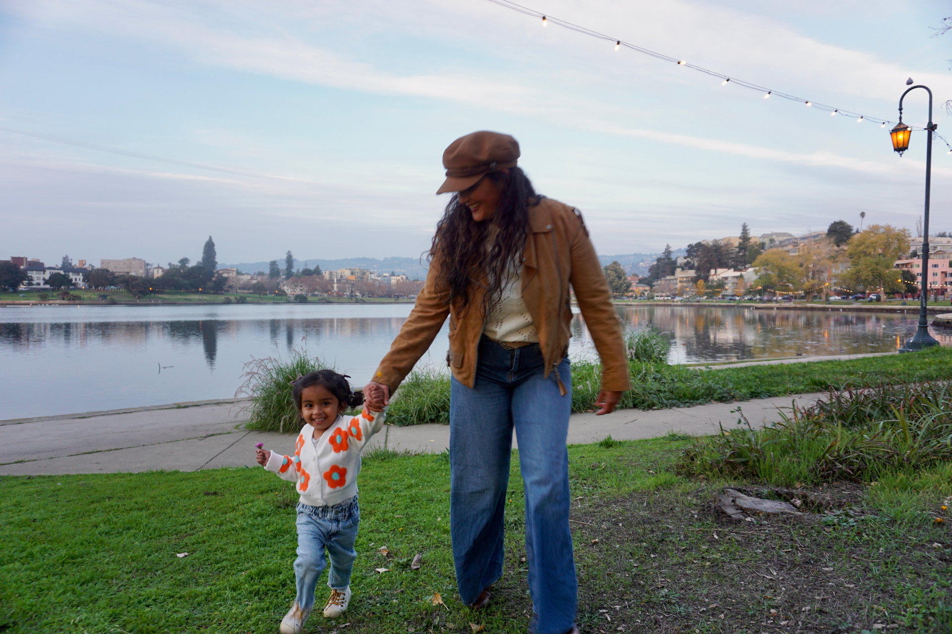 The Ramachandran mother and daughter holding hands and walking along the grass beside Lake Merritt at dusk