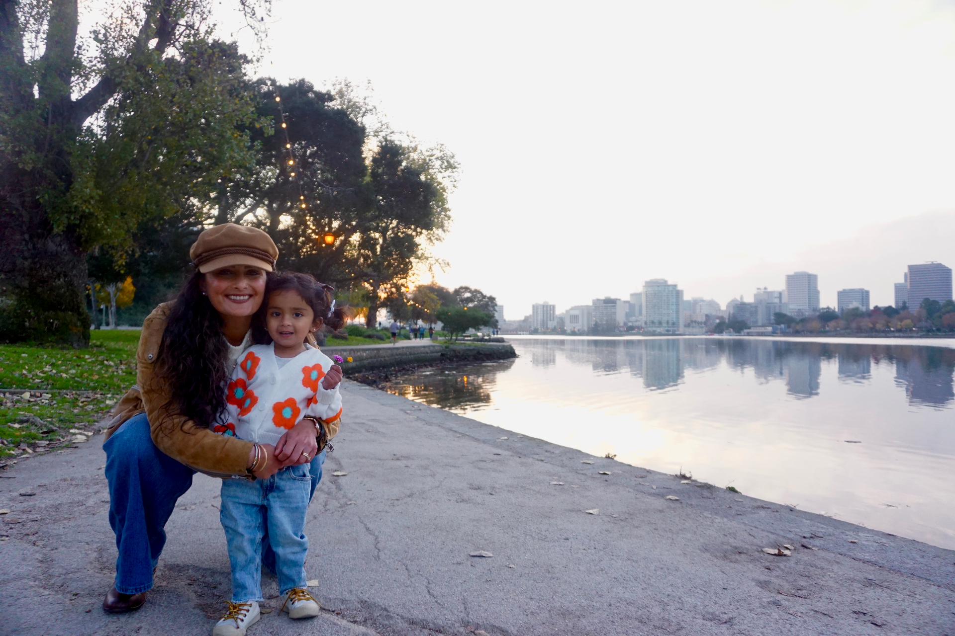The Ramachandran mother kneeling and hugging her daughter along the Lake Merritt path with the Oakland skyline at sunset