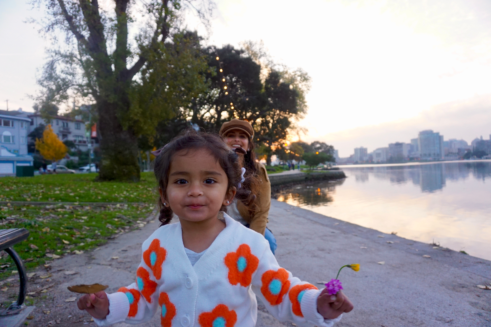 The Ramachandran daughter smiling at the camera holding a leaf and a flower with her mother laughing behind her by Lake Merritt