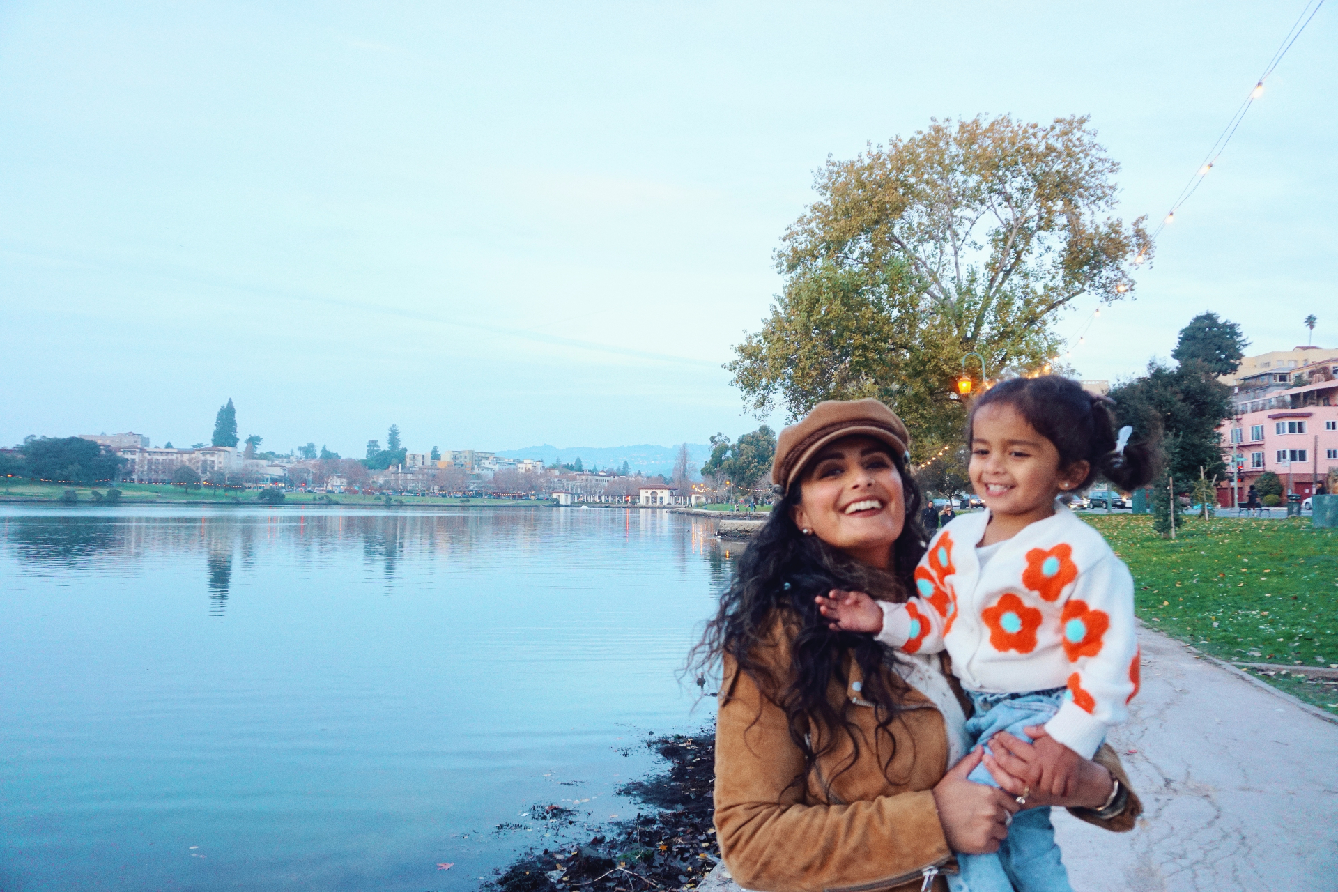 The Ramachandran mother holding her daughter by the edge of Lake Merritt at dusk with the shoreline lights reflecting on the water