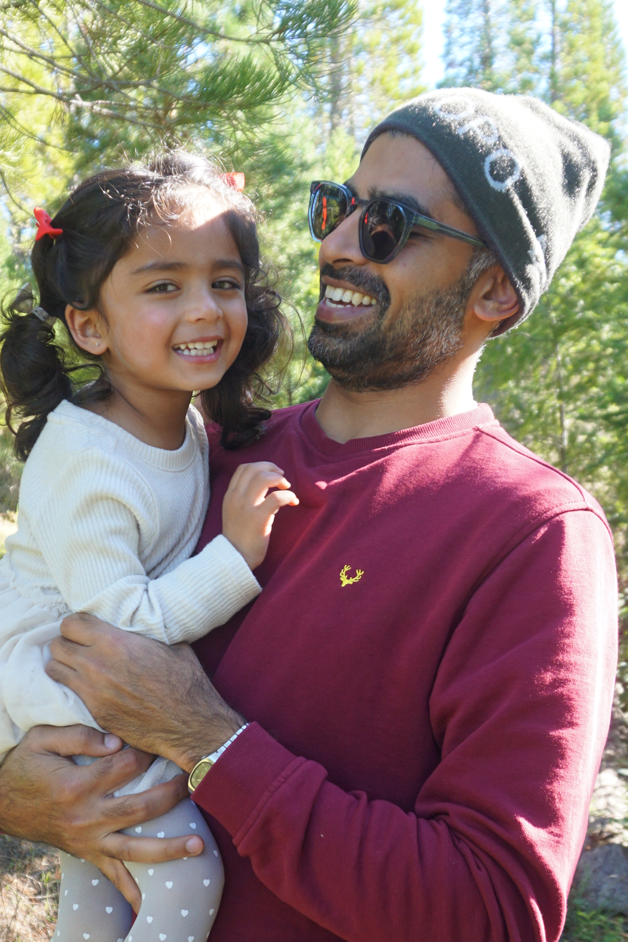 A joyful close-up of the Ramachandran father laughing while holding his smiling daughter in a sunlit pine forest