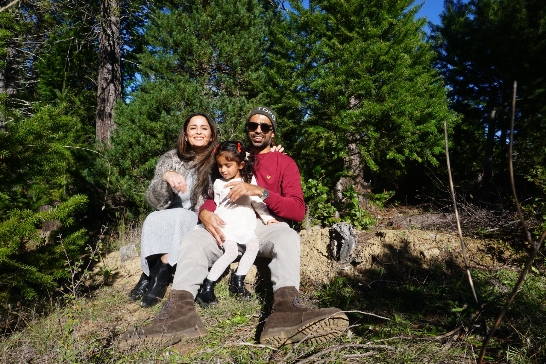 The Ramachandran family sitting together on a log in a sunlit pine forest with their daughter on their laps
