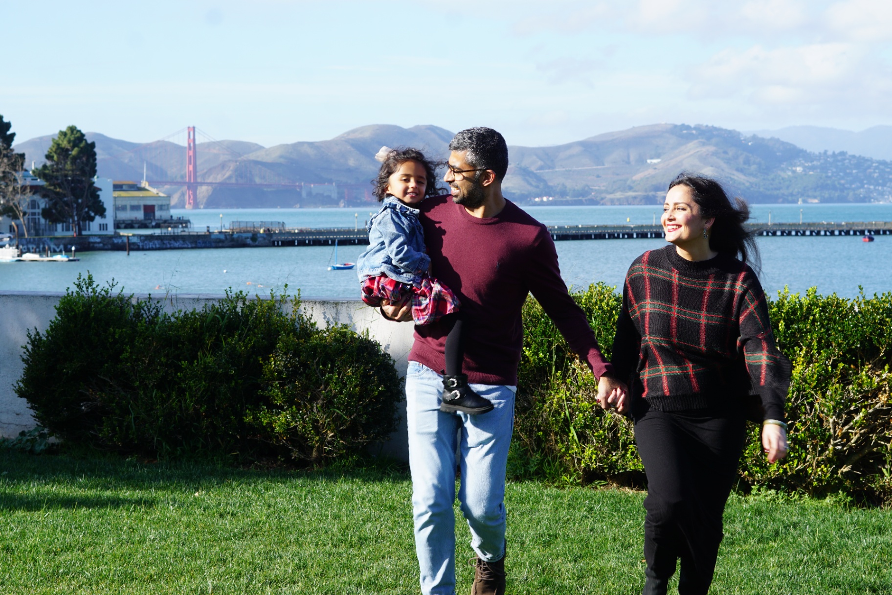 The Ramachandran family walking hand-in-hand on the grass, father holding their daughter, with the Golden Gate Bridge and bay in the background