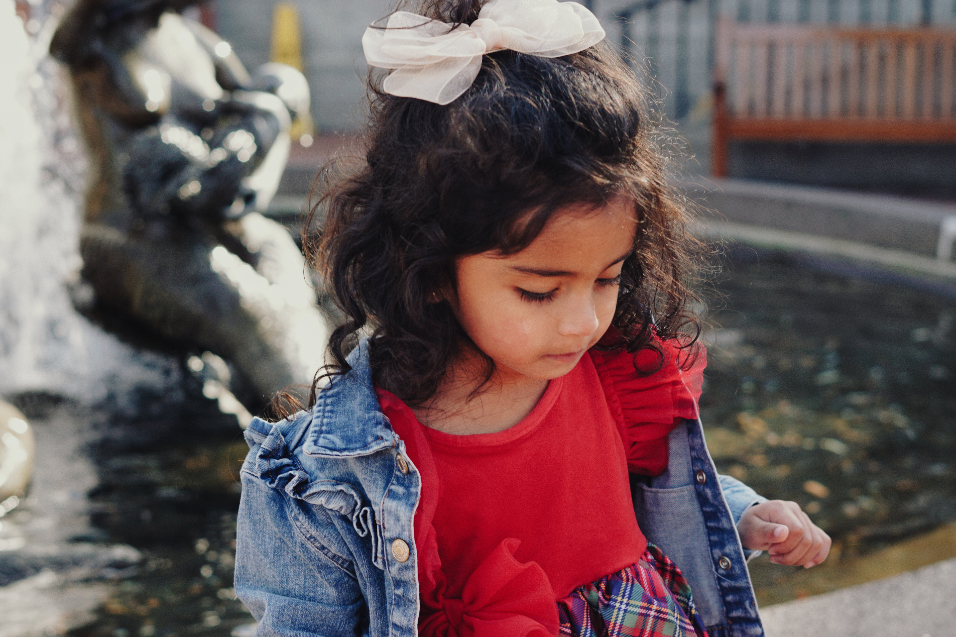 The Ramachandran daughter in a red dress and denim jacket with a bow in her hair, looking down beside a fountain