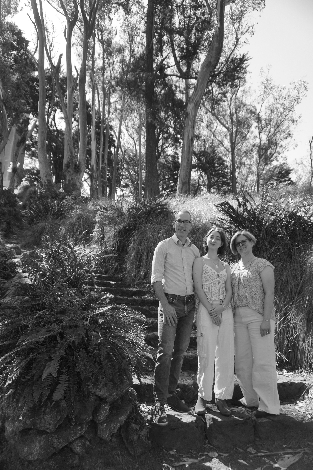 Black and white portrait of The Trimble family standing on stone steps surrounded by ferns and eucalyptus trees