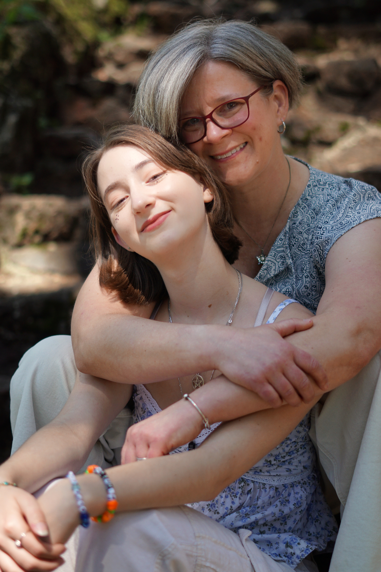 The Trimble mother hugging her daughter from behind on sunlit stone steps in the woods